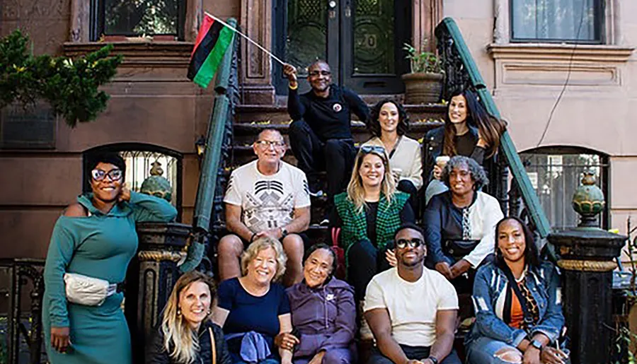 A diverse group of people happily posing together on the steps of a brownstone building, some sitting and others standing, with one person holding a flag.