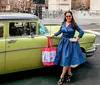 A smiling woman in a blue vintage-style dress is standing next to a classic green and yellow car while holding a shopping bag with city streets in the background