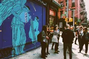 A group of people are standing on a city street corner in front of a striking blue building mural that features an angelic figure.