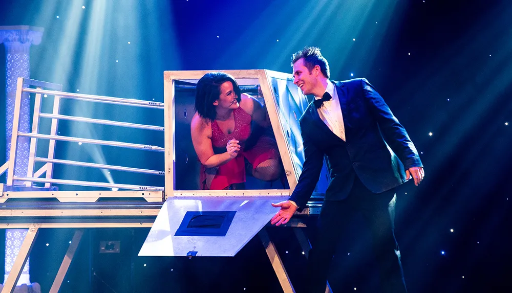 A magician in a tuxedo interacts with a smiling assistant emerging from a box on a stage with theatrical lighting
