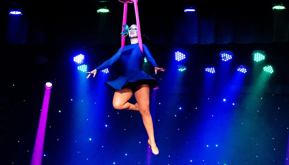 A performer is suspended in mid-air by aerial silks against a backdrop of vibrant stage lights