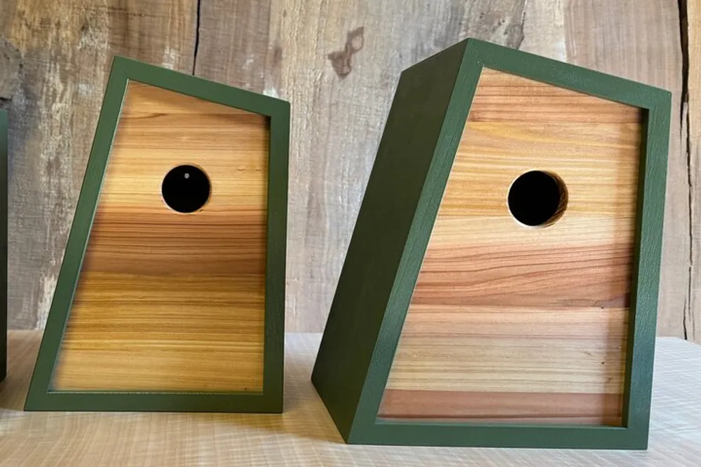 Two wooden birdhouses with green frames and contrasting natural-toned panels are displayed against a rustic wooden backdrop