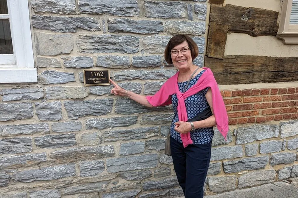 A smiling woman is pointing to a historical marker on a stone wall that indicates the year 1762