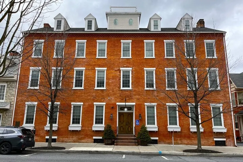 The image shows a large symmetrical brick building with a central entrance several windows on each floor and a decorative white trim under a cloudy sky