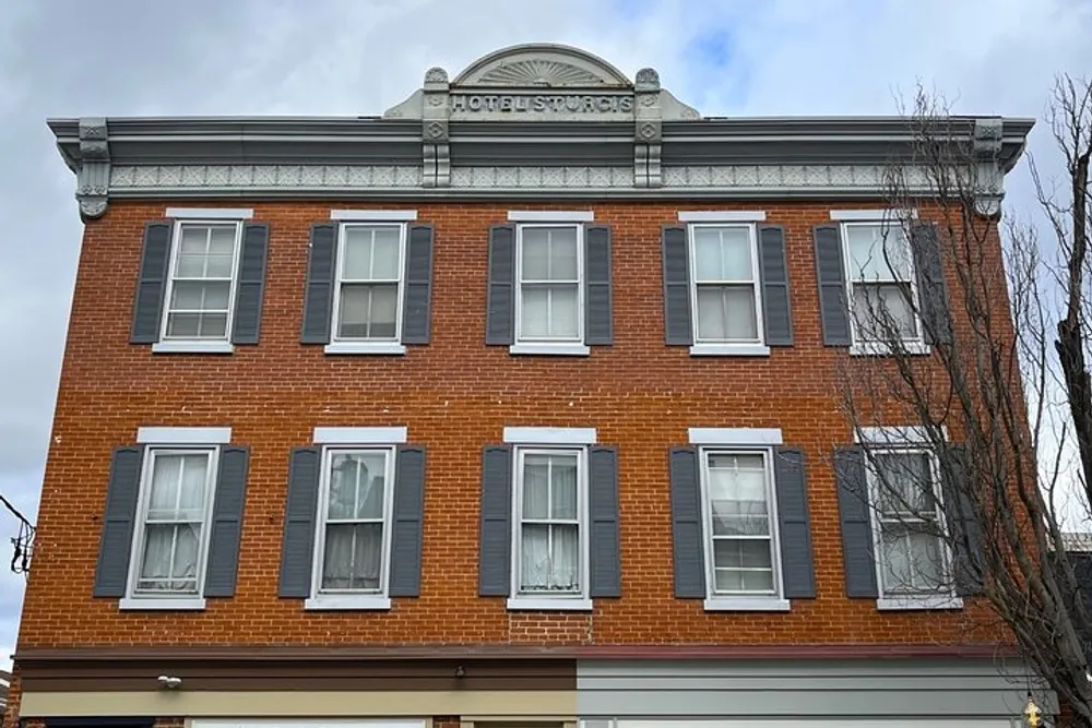 The image shows the upper facade of a classic red brick building with a sign that reads HOTEL STUDIOS under a decorative pediment featuring eight symmetrical windows with dark shutters against a cloudy sky