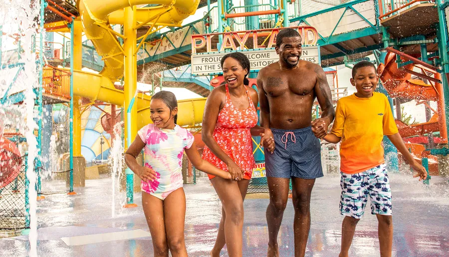 A family is joyfully running through water at a colorful water park with slides in the background.