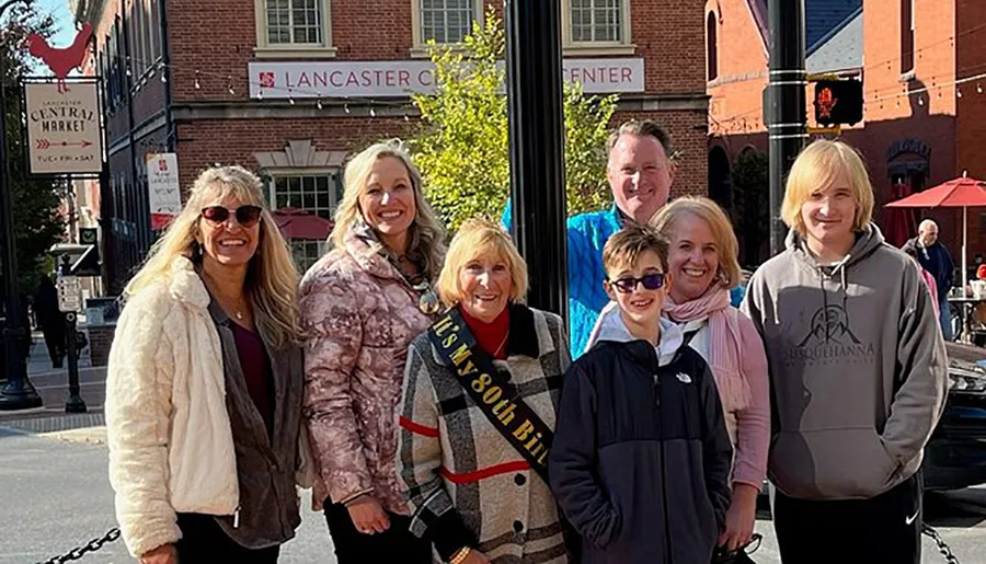 A group of seven people, including a woman wearing a sash that says It's My 80th Birthday, are smiling for a photo outdoors with a building labeled Lancaster Central Market in the background.