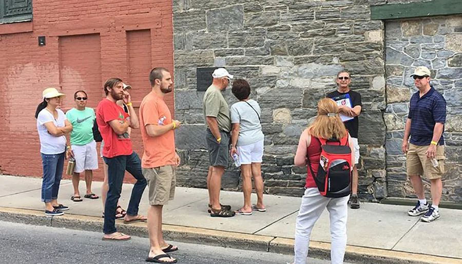 A group of people are standing outdoors on a sidewalk, seemingly listening or waiting, with some engaged in conversation and others looking in various directions.