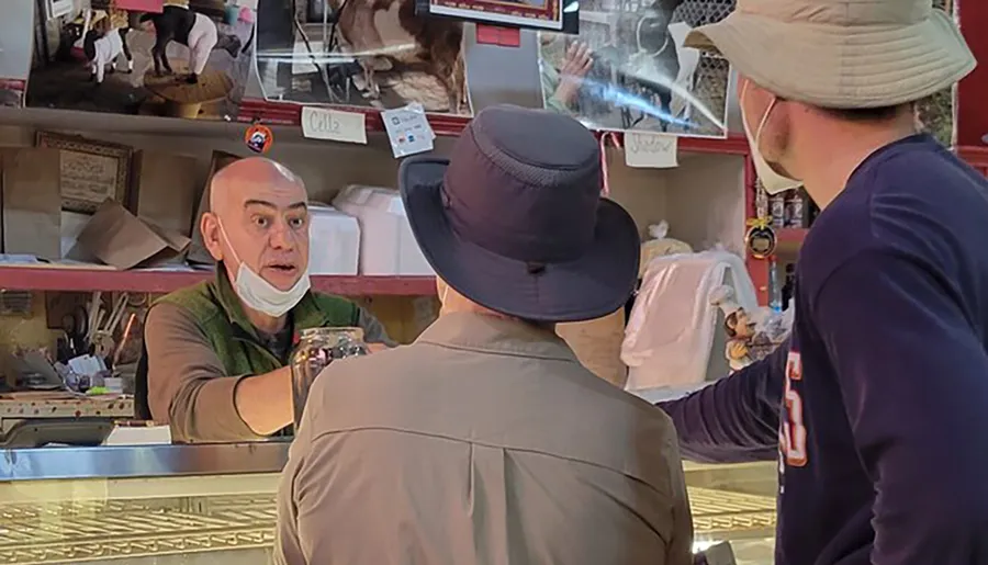 An employee at a food counter appears surprised or animated during an interaction with two customers wearing hats.