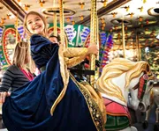 A joyful child in a princess costume is riding a carousel horse, smiling back at the camera.