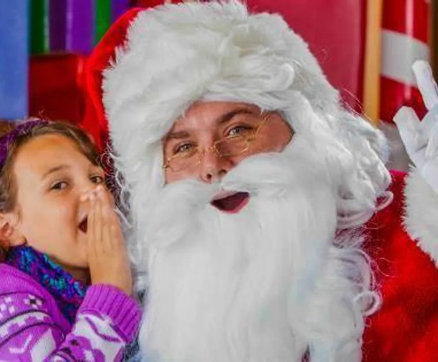 A person dressed as Santa Claus is winking and waving, while a child whispers into his ear, against a background of vibrant Christmas colors.