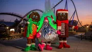 Three people in festive candy-themed costumes stand in front of a roller coaster at dusk.