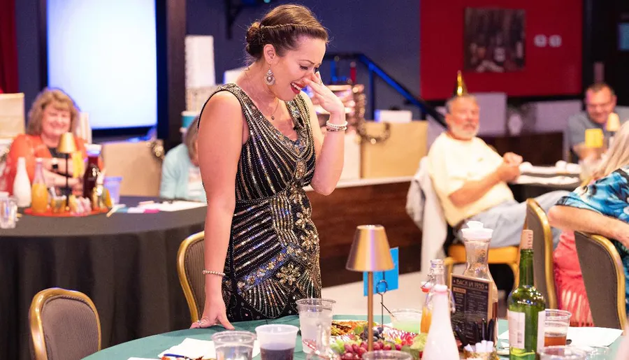 A woman in a sequined dress laughs heartily at a table during what appears to be a festive event or party.