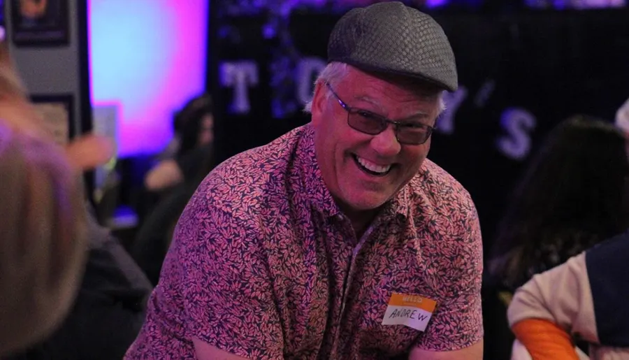 A man wearing a patterned shirt and a flat cap is smiling broadly with a name tag on his shirt in a room with purple lighting in the background.