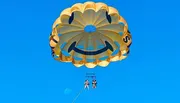 Two people are parasailing under a smiling face-decorated parachute against a clear blue sky.