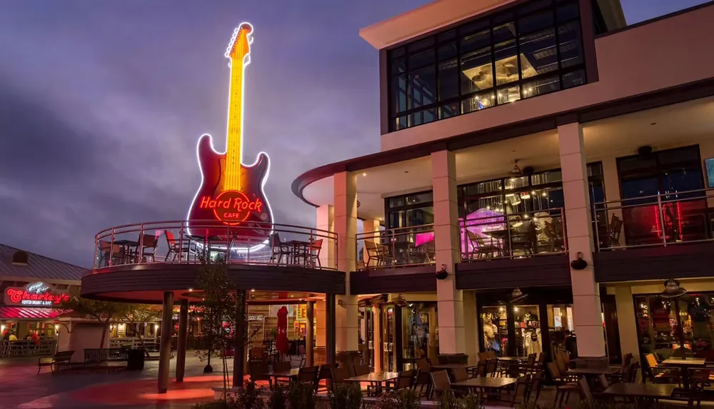 This is an evening photo of the Hard Rock Cafe with its iconic neon guitar sign situated in a lively outdoor shopping and dining area