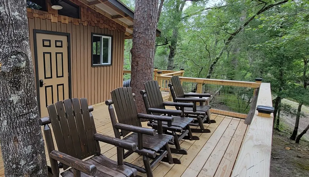 A cozy wooden cabin with a deck featuring several Adirondack chairs overlooks a tranquil forested area