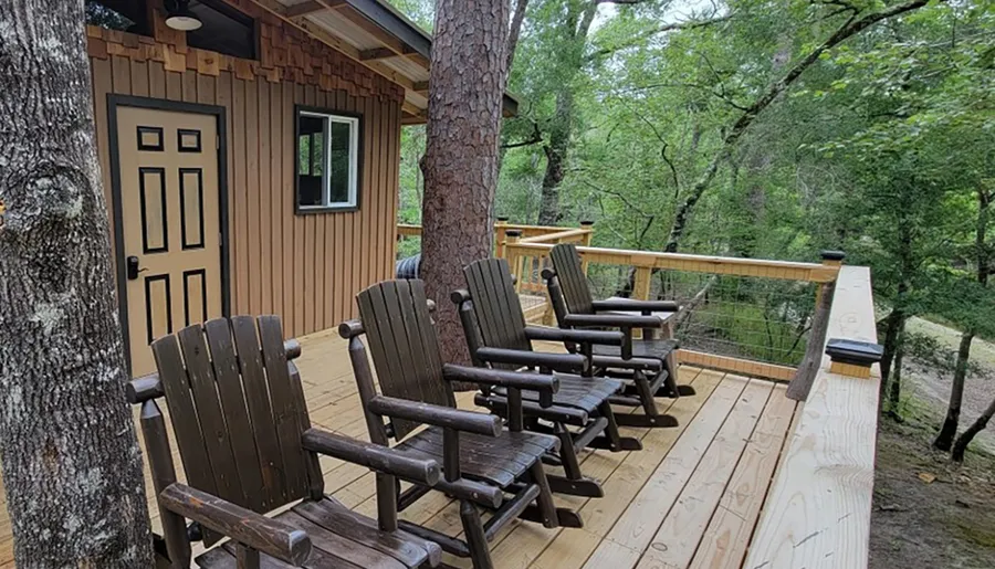 A cozy wooden cabin with a deck featuring several Adirondack chairs overlooks a tranquil forested area.