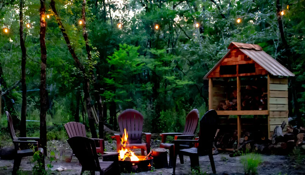 The image shows an inviting outdoor evening scene with a crackling fire pit Adirondack chairs hanging lights and a woodshed set amongst verdant trees