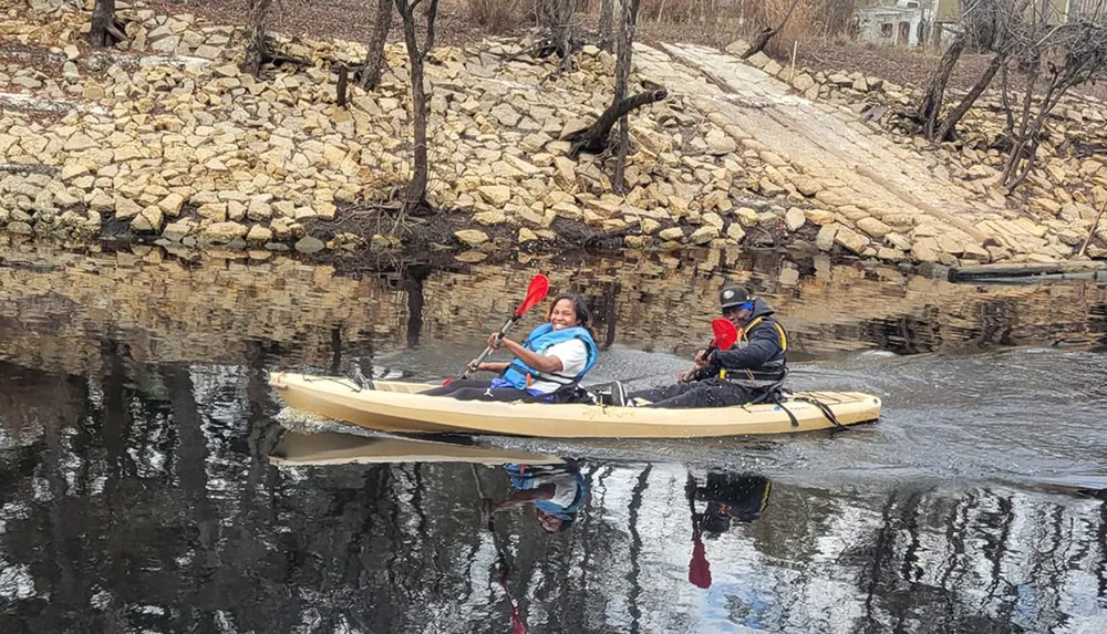 Two people are smiling and kayaking together on a calm river with a rocky riverbank in the background