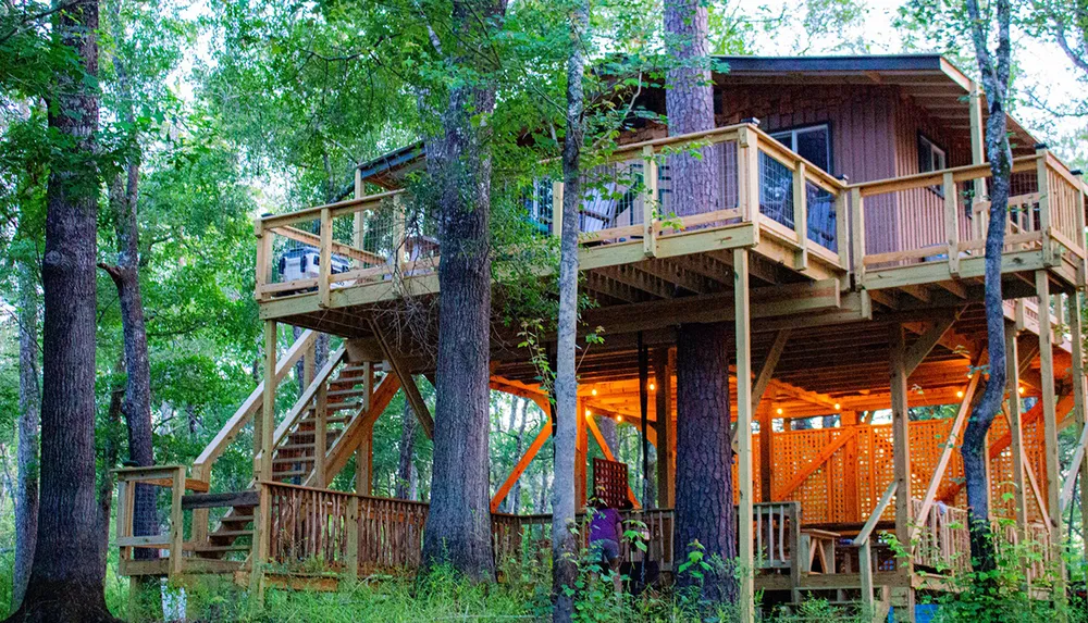 The image shows a warm inviting treehouse nestled among trees with a staircase and multiple levels of wooden decking