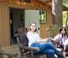 Two people are smiling and sitting on wooden chairs on the porch of a cabin surrounded by trees