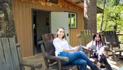 Two people are smiling and sitting on wooden chairs on the porch of a cabin, surrounded by trees.