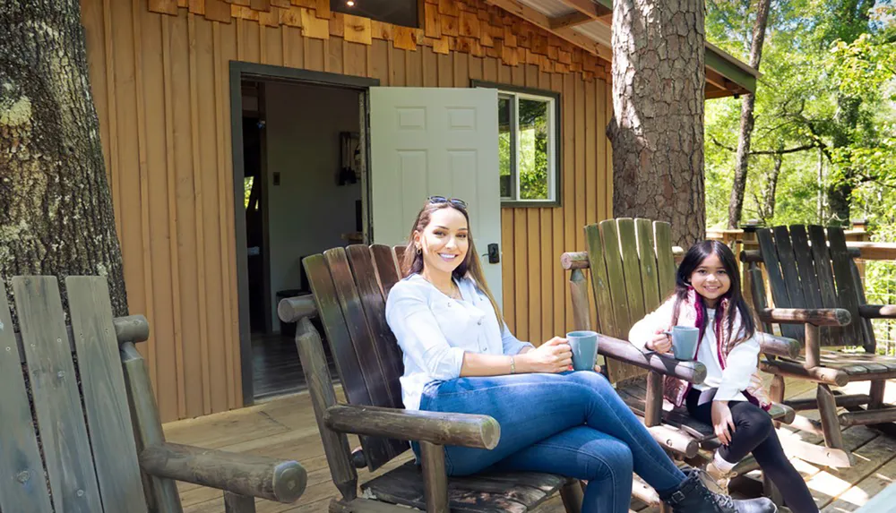 Two people are smiling and sitting on wooden chairs on the porch of a cabin surrounded by trees