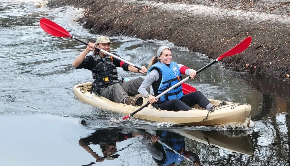 Two people are paddling in a kayak on a calm river with reflections visible in the water