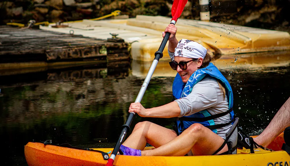 A person wearing a blue life jacket and sunglasses is enthusiastically paddling a yellow kayak with water droplets visible in the air