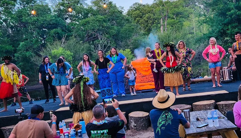 An audience watches and captures a moment on camera as a diverse group of people some in colorful attire participate in a festive outdoor event with a stage set in a natural wooded area