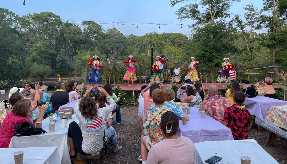 A group of people is watching a vibrant outdoor performance with dancers in colorful costumes on stage under string lights