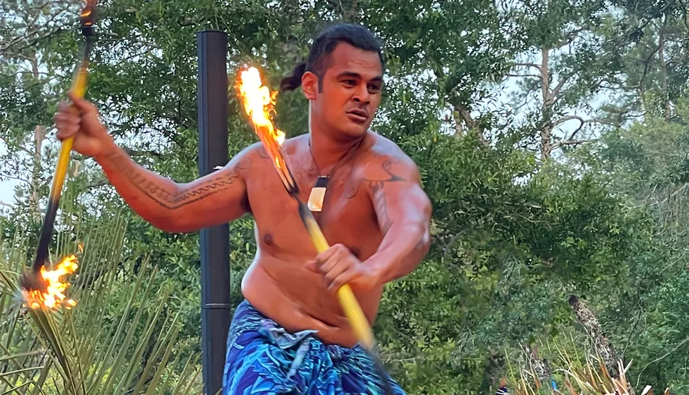 A shirtless performer with traditional tattoos wields flaming sticks possibly as part of a cultural or fire dance performance set against a backdrop of lush greenery