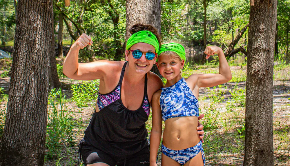 A woman and a young child are smiling and striking playful strong-arm poses in a sunny forest setting