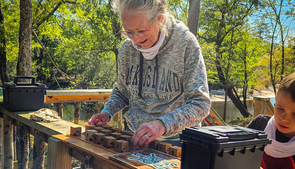 A woman is interacting with a set of blocks on a table outdoors in a sunny wooded area while a child looks on with interest