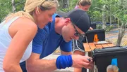 Two people are closely examining an object on top of a set of black boxes labeled with numbers, suggesting they might be engaged in some kind of outdoor activity or challenge.