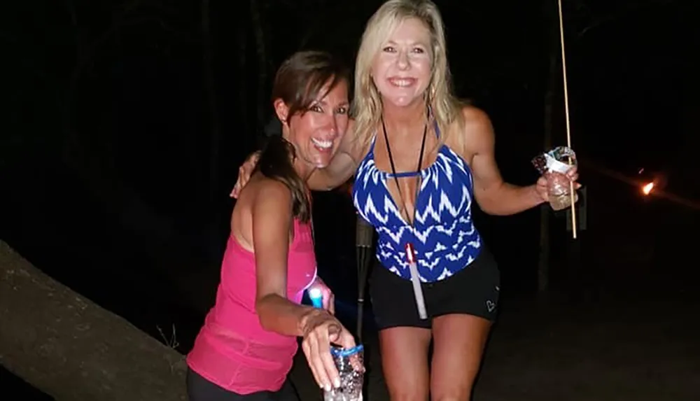 Two women are posing with smiles at night one of which is holding a marshmallow on a stick and a flashlight indicating they might be enjoying some outdoor activities such as camping or a bonfire