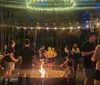 People are gathered around a campfire at twilight enjoying conversation and roasting marshmallows under a canopy of string lights