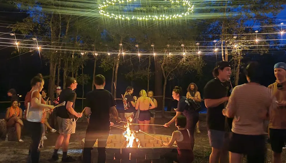 People are gathered around a campfire at twilight enjoying conversation and roasting marshmallows under a canopy of string lights