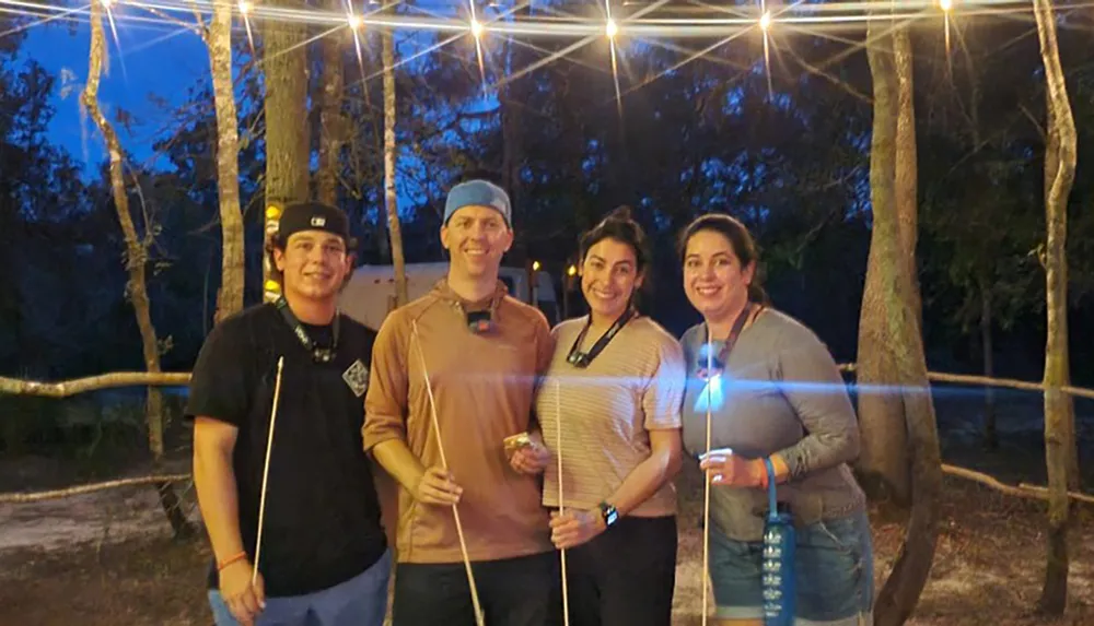 Four people are smiling for a photo at dusk with string lights overhead two of them holding marshmallow roasting sticks