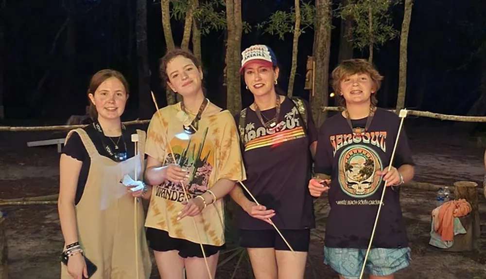 Four individuals are smiling at the camera during a nighttime outdoor activity holding roasting sticks with marshmallows