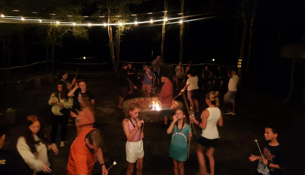 A group of people are enjoying an outdoor gathering at night around a fire pit some roasting marshmallows under string lights