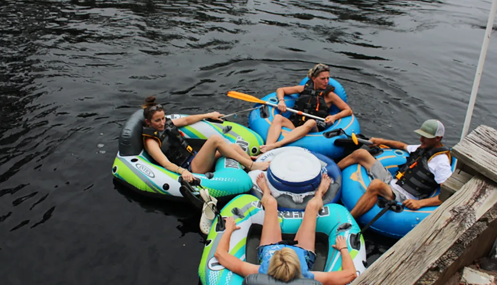 A group of people in life vests are lounging and holding paddles while sitting in linked inflatable tubes on a calm body of water