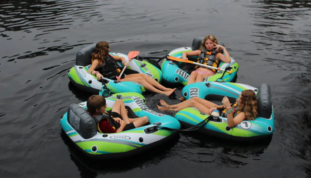 Four individuals are floating on interconnected inflatable rafts on calm water each holding a paddle