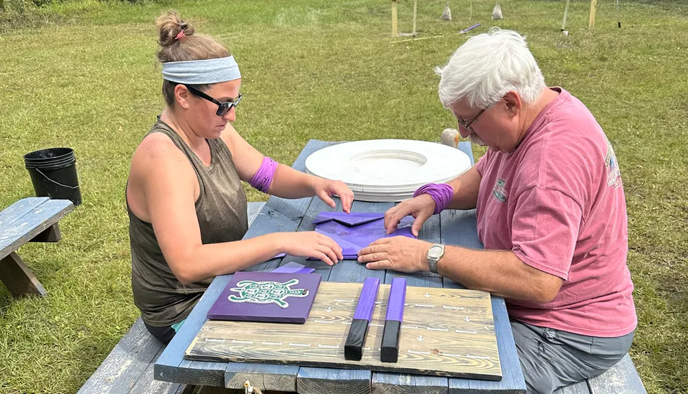 Two individuals are engaged in a craft activity at a picnic table handling painted wooden pieces and stencils