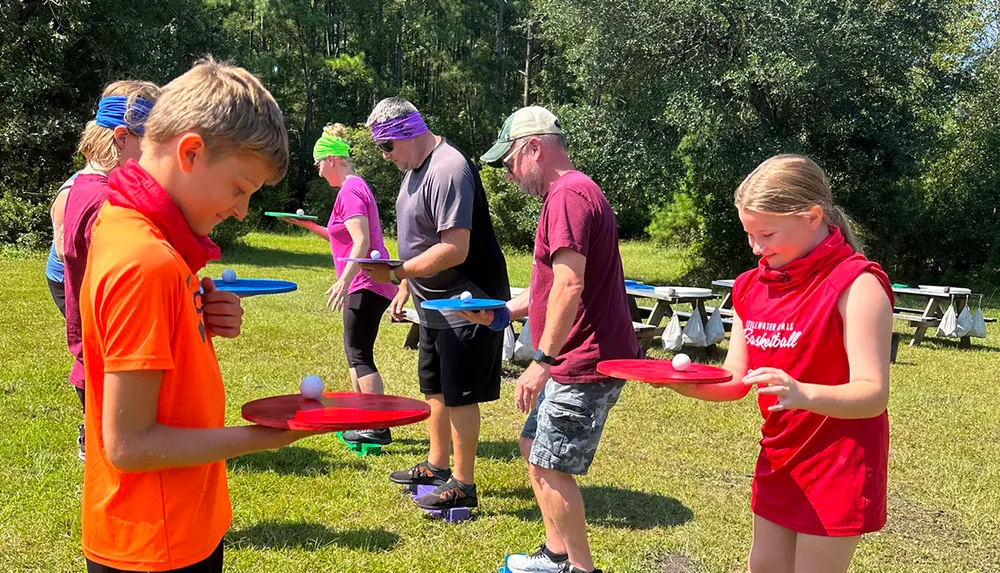 A group of people including children and adults are concentrating on balancing balls on plates outdoors on a sunny day