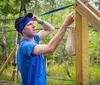 A person in a blue shirt and headband appears to be engaged in an outdoor physical activity or construction task wiping sweat from their forehead near a wooden structure with hanging ropes or straps