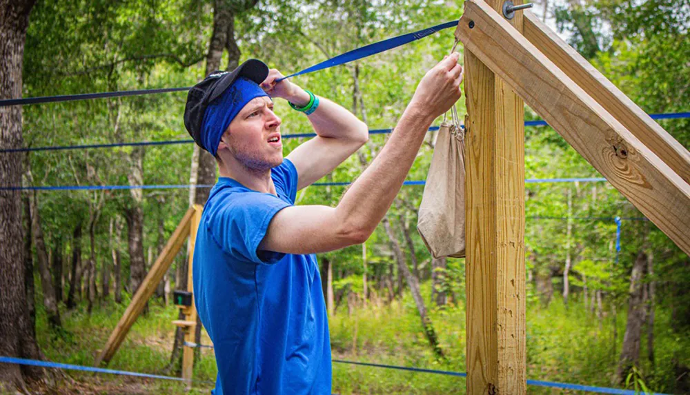 A person in a blue shirt and headband appears to be engaged in an outdoor physical activity or construction task wiping sweat from their forehead near a wooden structure with hanging ropes or straps