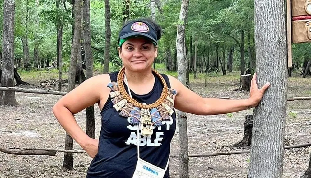 A smiling person stands in a forested area wearing a cap a necklace with large elements and a tank top with the word ABLE while touching a tree trunk