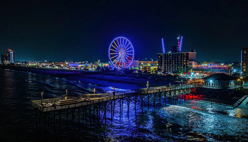 A brightly lit ferris wheel dominates the night skyline by a pier along a bustling beachfront cityscape